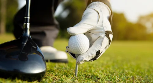 A close up on the golf course of a golfer leaning down, wearing a golf glove, teeing up a golf ball with their driver next to the tee.