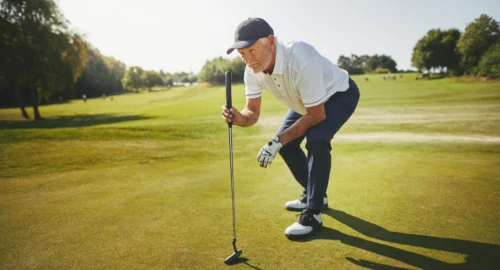 A golfer bending his legs, leaning on a golf putter on the golf course to see where his golf ball is heading toward the hole.