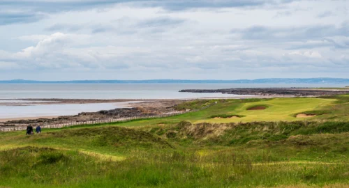 A links golf course, featuring long grass, no trees, and a coastal view.