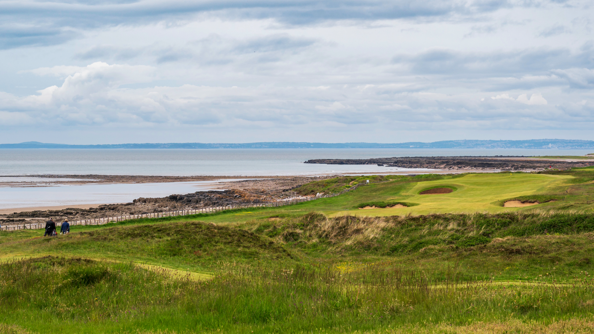 A links golf course, featuring long grass, no trees, and a coastal view.