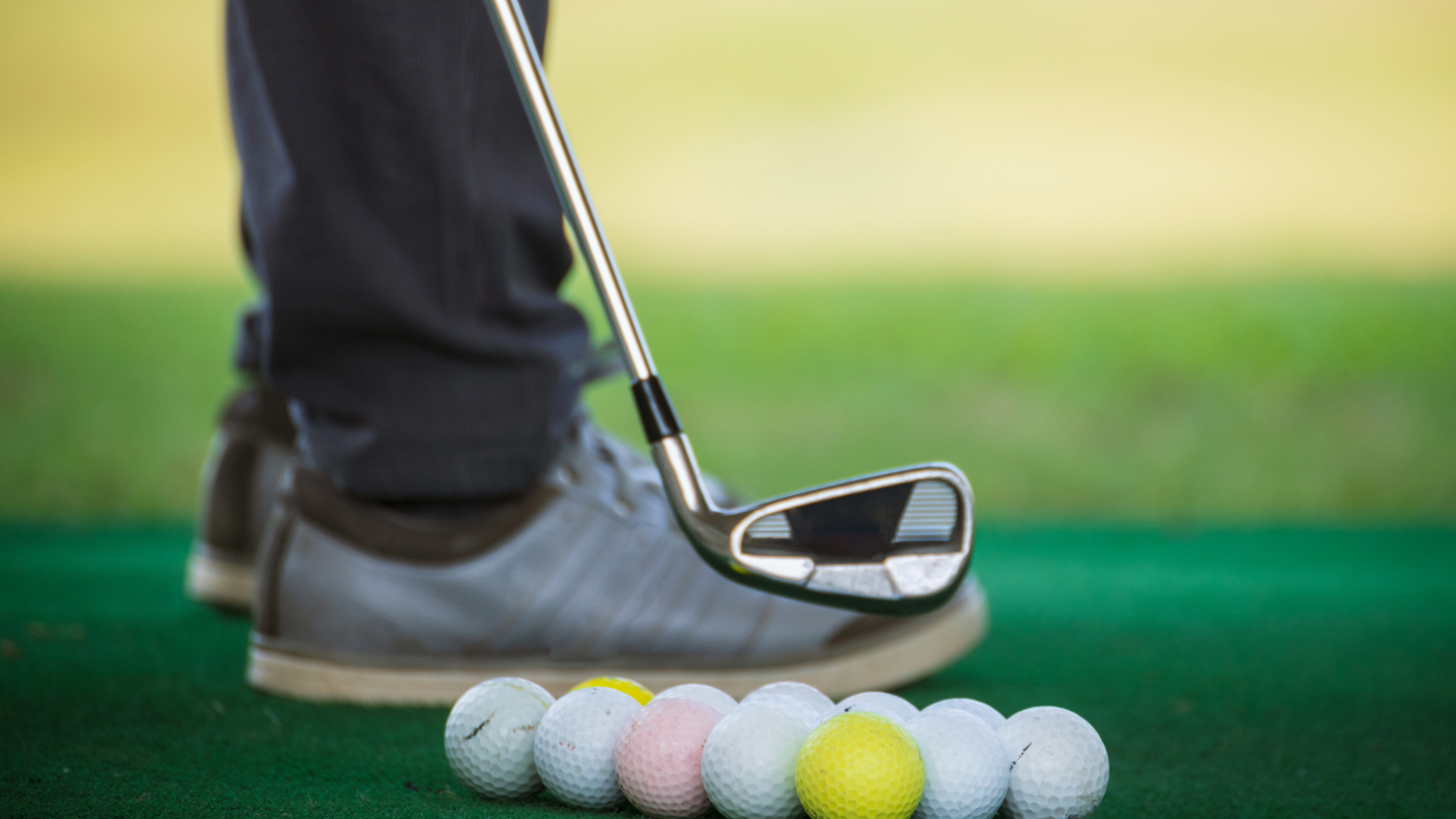 Close up of a golfer's shoes, with many golf balls and a golf club in the foreground.