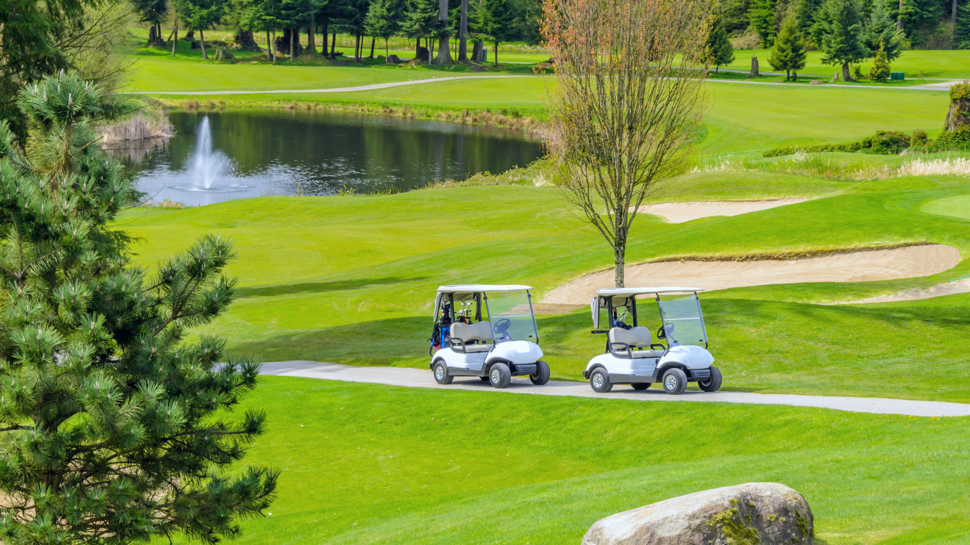 Two golf carts on the golf course cart track, with trees, water features, bunkers and lots of grass.